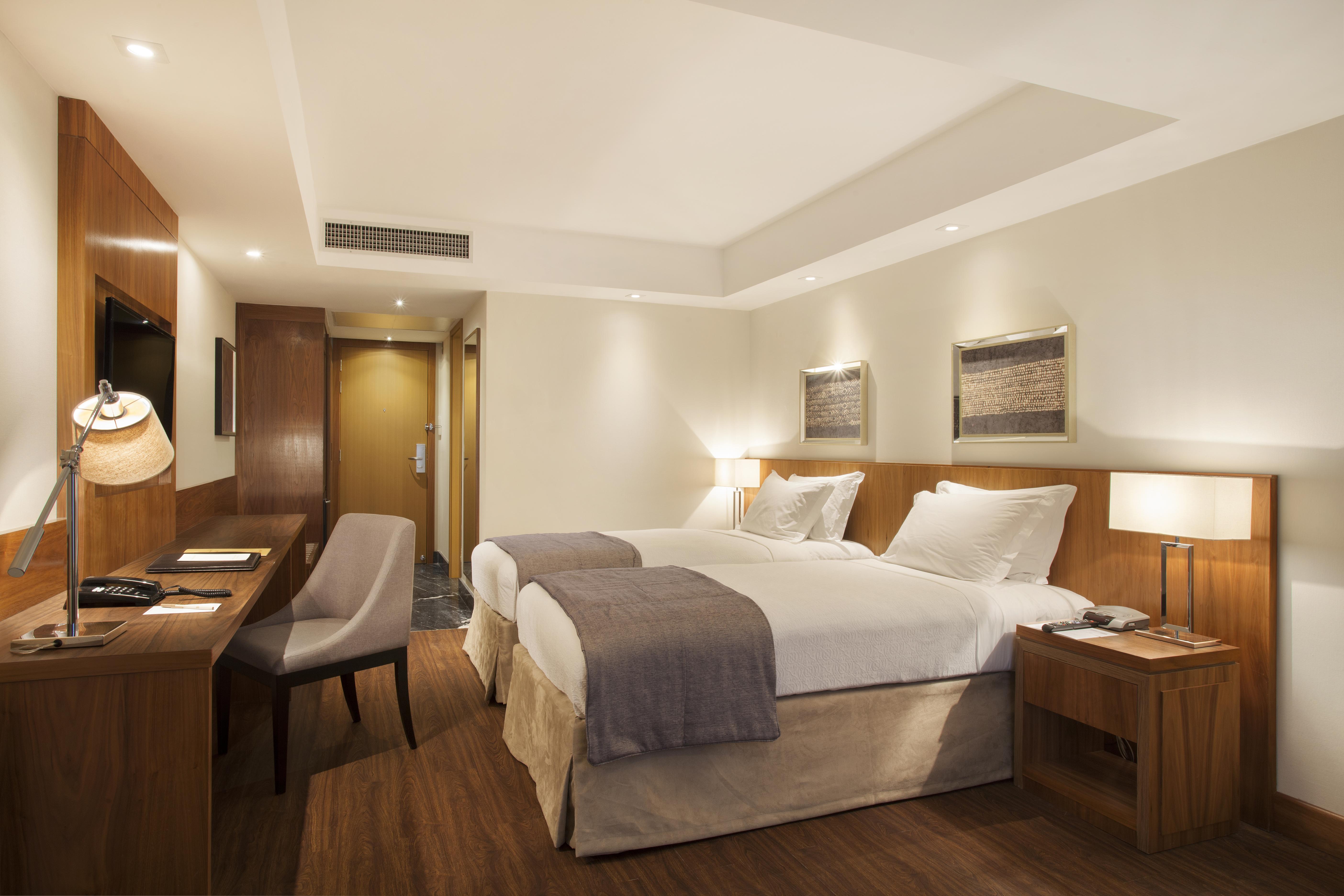 ホテル ウィンザー マラペンディ リオデジャネイロ エクステリア 写真 The photo displays a modern hotel room featuring two twin beds with crisp white linens and gray throw blankets. The room has a clean and minimalist design, with wooden accents, including a bedside table and a desk. To the left, there's a small desk a