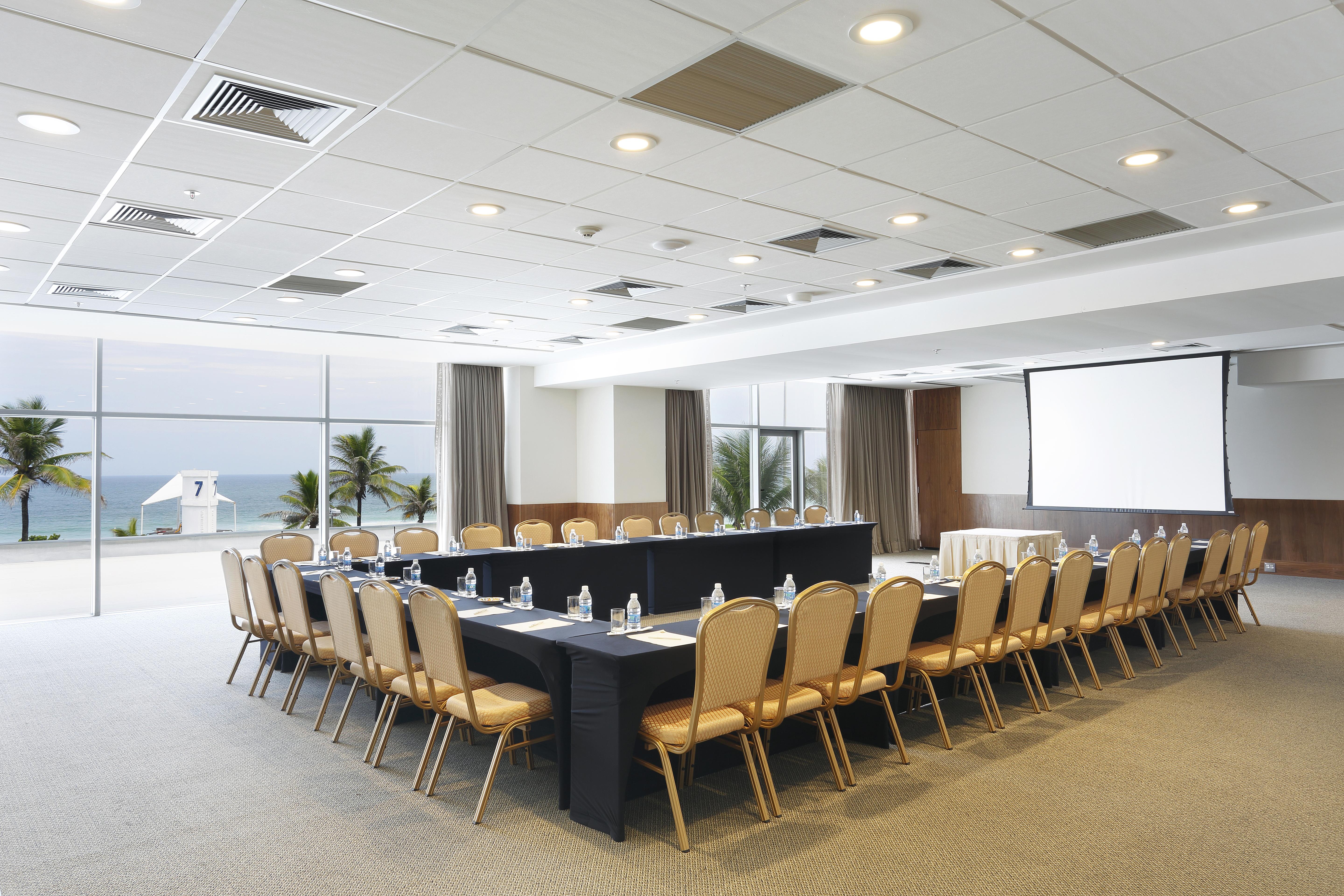 ホテル ウィンザー マラペンディ リオデジャネイロ エクステリア 写真 The photo shows a spacious conference room set up for a meeting. It features a long, rectangular table surrounded by a number of beige chairs. At the front, there is a large screen, likely for presentations. The room has large windows that provide a 